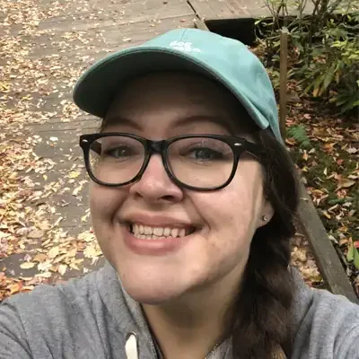 Close-up outdoor selfie of a person wearing black-framed glasses, a teal baseball cap, and a gray hoodie.