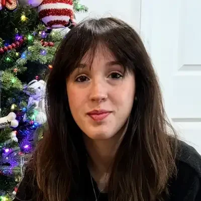 Close-up portrait of a person with long dark hair wearing a black top, seated indoors in front of a decorated Christmas tree with colorful lights and ornaments.